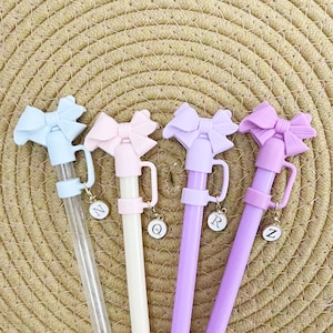 May include: Four pastel-colored pens with bow-shaped tops. Each pen has a different color: light blue, pink, lavender, and purple. Each pen has a charm with a letter. The pens are on a woven background.