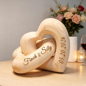 May include: A light-colored wooden sculpture consisting of two interlocking heart shapes. The wood features a visible grain pattern. The text "Finch & Sily" is engraved on the front heart, and the date "05.20.2026" is engraved on the side.
