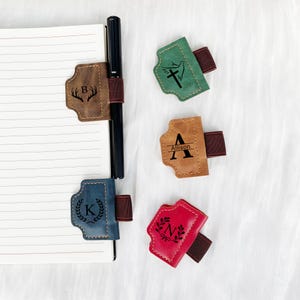 Könnte beinhalten: Ein Stifthalter mit einem braunen Lederriemen, der an einem Notizbuch befestigt ist. Der Riemen ist mit dem Buchstaben "B" und einem Geweih personalisiert. Zusätzliche Stifthalter in Grün, Beige und Rot sind ebenfalls abgebildet, jeder mit einem anderen Design und Initialen.
