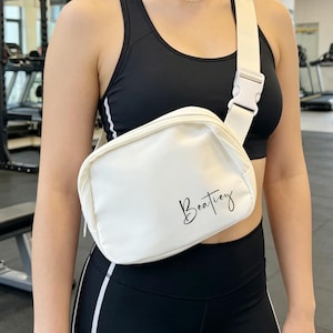 May include: A white fanny pack with a black script that reads "Beatify" is worn across the body. The bag has a white strap and a white plastic buckle. The person is wearing a black sports bra and black leggings.