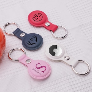 May include: Four leather AirTag keyrings in pink, white, navy, and red. Each keyring has a silver ring and a snap closure. The pink keyring has the letter "S" and the name "Salvin" printed on it.