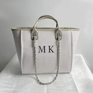 Puede incluir: Un bolso tote de lona blanco con una correa de cadena plateada y las iniciales "MK" bordadas en la parte delantera.
