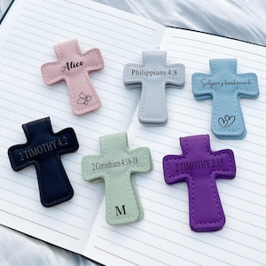 May include: A collection of cross-shaped bookmarks in various colors, including pink, gray, blue, black, green, and purple. Each bookmark features text, such as Bible verses or names. Some have decorative elements like a butterfly or a heart.