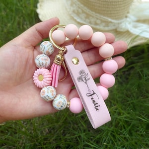 May include: A circular keychain bracelet featuring smooth light pink silicone beads, white beads with orange floral patterns, and a pink daisy-shaped bead. It includes a gold ring, a pink tassel, and a pink leather strap engraved with roses and the name Featie.