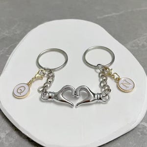 May include: Two silver keyrings linked by a heart-shaped clasp. Each keyring has a circular charm with a letter inside a white circle. One charm has the letter "Q" and the other has the letter "N".