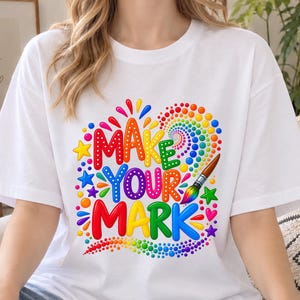 May include: A white short-sleeved t-shirt featuring a colorful graphic design with the text "MAKE YOUR MARK" in bold, multi-colored letters. The design includes a paintbrush, small stars, hearts, and a swirling pattern of multi-colored dots.