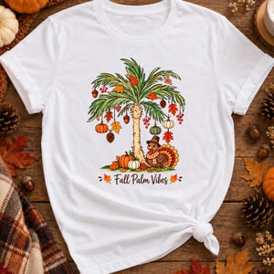 May include: A white short-sleeved t-shirt featuring a graphic of a palm tree decorated with pumpkins, acorns, and autumn leaves. A turkey wearing a pilgrim hat stands at the base of the tree. The text "Fall Palm Vibes" is printed below.