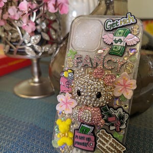 K&ouml;nnte beinhalten: Eine transparente Handyh&uuml;lle, verziert mit Strasssteinen, Glitzer und bunten Aufklebern. Die Aufkleber zeigen einen Hello Kitty-Charakter, einen Geldbeutel, eine rosa Blume und den Text "Get rich", "Fancy" und "Mach dir keine Sorgen um Dinge, die du nicht kontrollieren kannst".