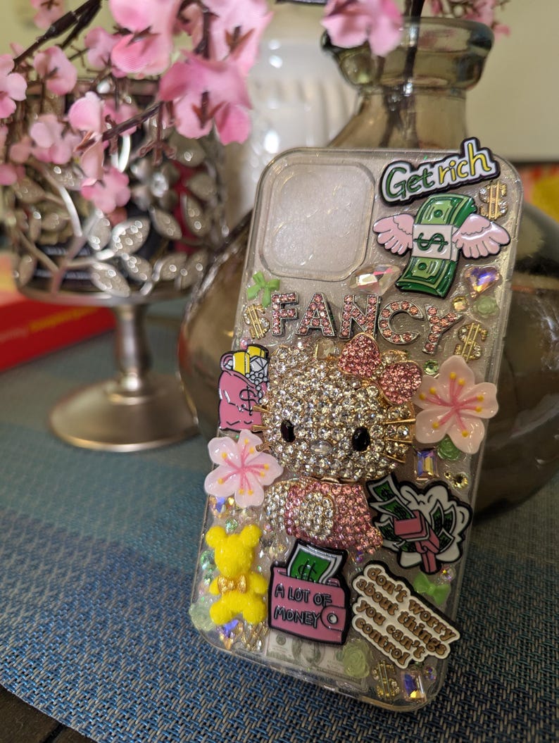 K&ouml;nnte beinhalten: Eine transparente Handyh&uuml;lle, verziert mit Strasssteinen, Glitzer und bunten Aufklebern. Die Aufkleber zeigen einen Hello Kitty-Charakter, einen Geldbeutel, eine rosa Blume und den Text "Get rich", "Fancy" und "Mach dir keine Sorgen um Dinge, die du nicht kontrollieren kannst".