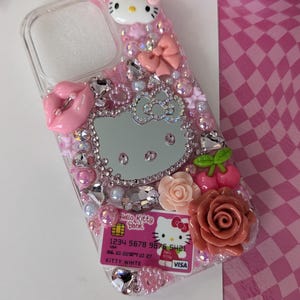 May include: A clear phone case decorated with pink Hello Kitty themed embellishments. The case features a Hello Kitty design, rhinestones, bows, and a decorative credit card. The case is adorned with pink roses and cherries.
