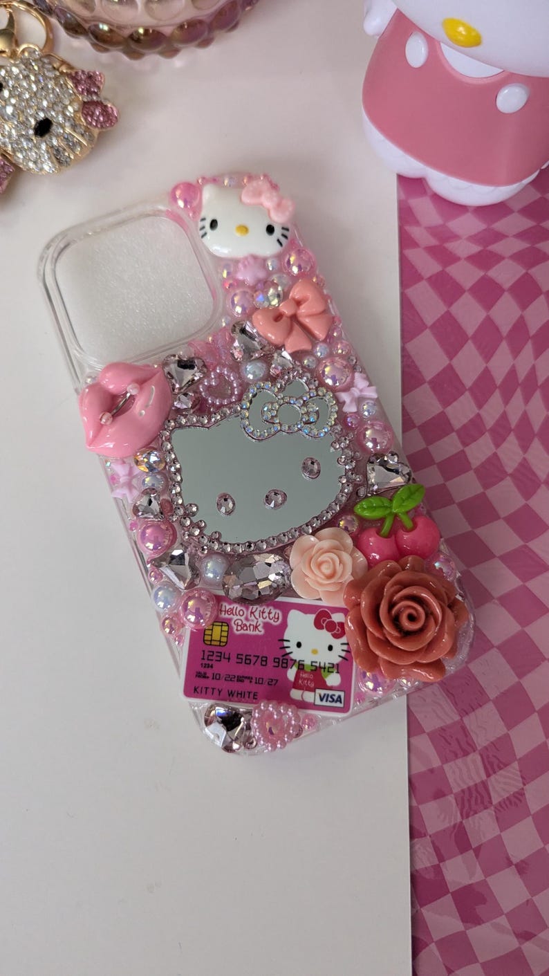 May include: A clear phone case decorated with pink Hello Kitty themed embellishments. The case features a Hello Kitty design, rhinestones, bows, and a decorative credit card. The case is adorned with pink roses and cherries.