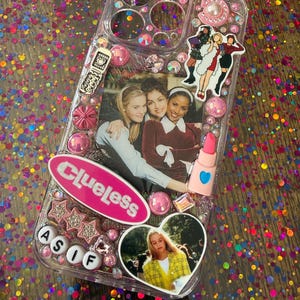 May include: Clear phone case decorated with pink and clear rhinestones, a pink bow, and a photo of the movie "Clueless". The case features a "Clueless" logo, star-shaped embellishments, and a lipstick charm.