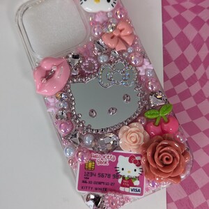 May include: A clear phone case decorated with pink rhinestones, bows, and Hello Kitty-themed embellishments. Features a Hello Kitty face, a mirrored Hello Kitty silhouette, and a Hello Kitty Bank credit card design. Includes a pink lip and rose accents.