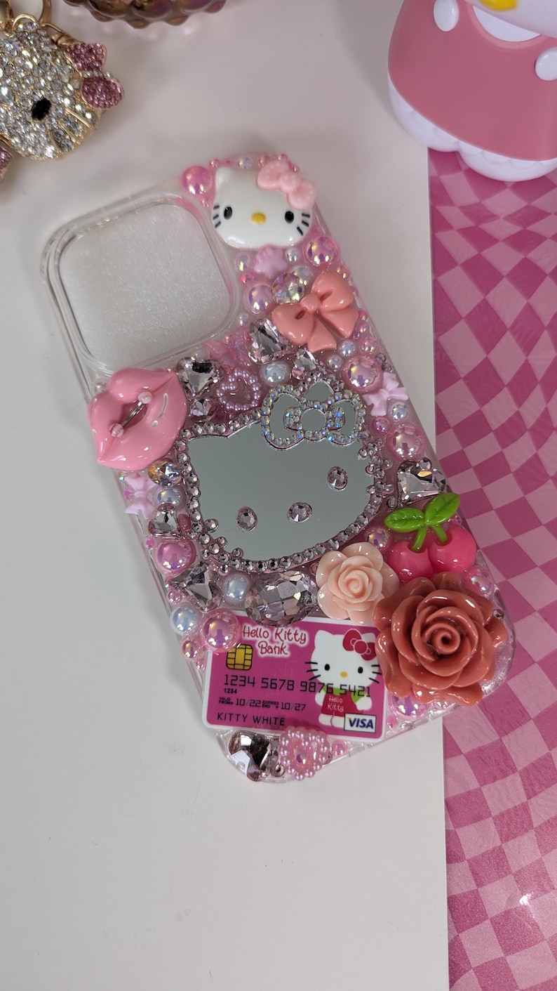 May include: A clear phone case decorated with pink rhinestones, bows, and Hello Kitty-themed embellishments. Features a Hello Kitty face, a mirrored Hello Kitty silhouette, and a Hello Kitty Bank credit card design. Includes a pink lip and rose accents.