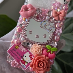 May include: A pink and clear phone case decorated with Hello Kitty, a mirror, pink lips, bows, roses, and rhinestones. The case also features a Hello Kitty Bank credit card design.