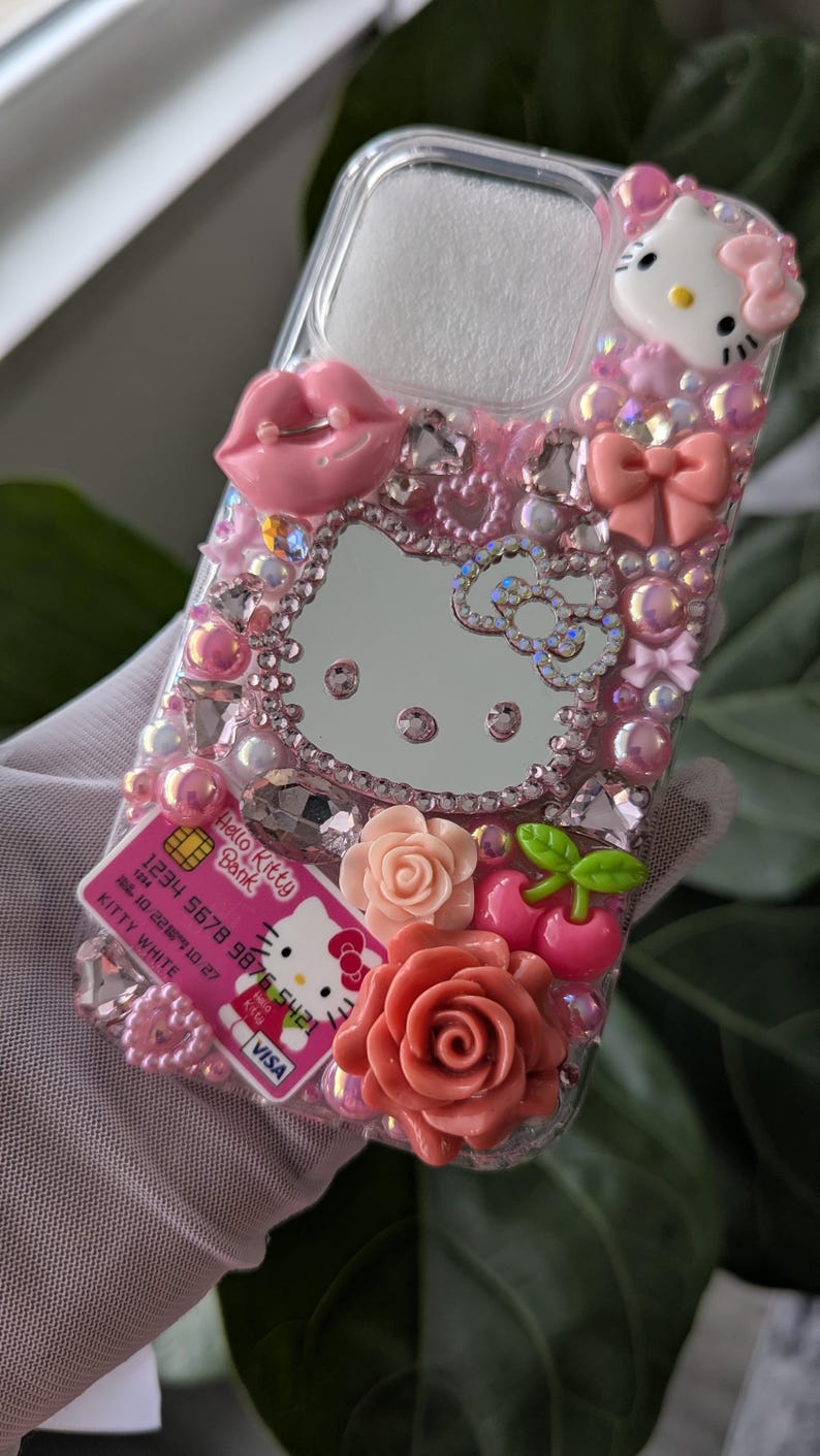 May include: A pink and clear phone case decorated with Hello Kitty, a mirror, pink lips, bows, roses, and rhinestones. The case also features a Hello Kitty Bank credit card design.