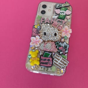K&ouml;nnte beinhalten: Eine transparente Handyh&uuml;lle, die mit Strasssteinen, Charms und Aufklebern verziert ist. Die H&uuml;lle zeigt ein Hello Kitty-Motiv, eine rosa Handtasche mit dem Text "A lot of money" sowie die Texte "Get rich" und "Don't worry about things you can't control".