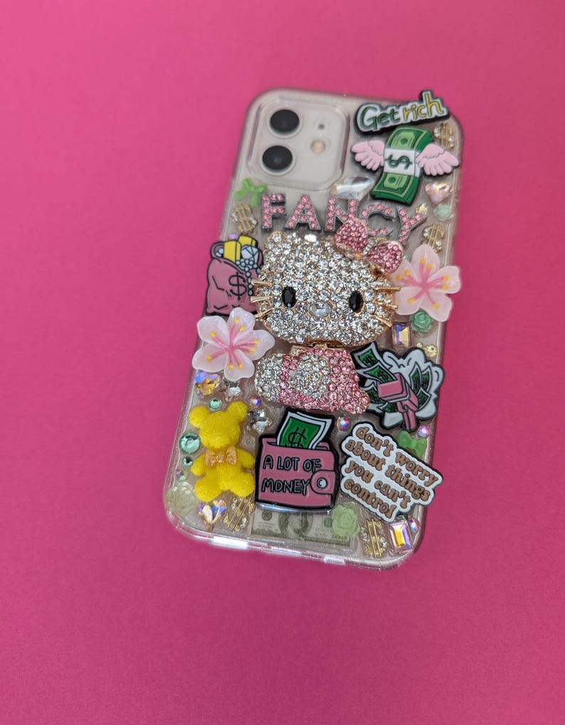 K&ouml;nnte beinhalten: Eine transparente Handyh&uuml;lle, die mit Strasssteinen, Charms und Aufklebern verziert ist. Die H&uuml;lle zeigt ein Hello Kitty-Motiv, eine rosa Handtasche mit dem Text "A lot of money" sowie die Texte "Get rich" und "Don't worry about things you can't control".