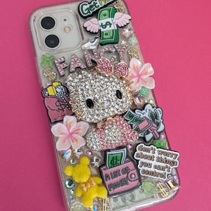 K&ouml;nnte beinhalten: Eine transparente Handyh&uuml;lle, die mit Strasssteinen, Glitzer und verschiedenen Charms verziert ist. Zu den Charms geh&ouml;ren ein Hello Kitty-Charakter, eine rosa Handtasche mit einem Dollarzeichen, ein gr&uuml;ner Schein mit Fl&uuml;geln, eine rosa und gr&uuml;ne Blume, ein gelber Teddyb&auml;r und eine rosa Geldb&ouml;rse mit dem Text "A lot of money". Die H&uuml;lle tr&auml;gt au&szlig;erdem den Text "Get rich" und "Fancy".