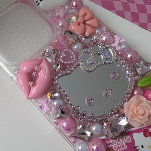May include: A clear phone case bedazzled with pink and clear rhinestones, pearls, and charms. The case features a Hello Kitty design, a pink bow, and a pink lip charm. A Hello Kitty Bank credit card is visible.