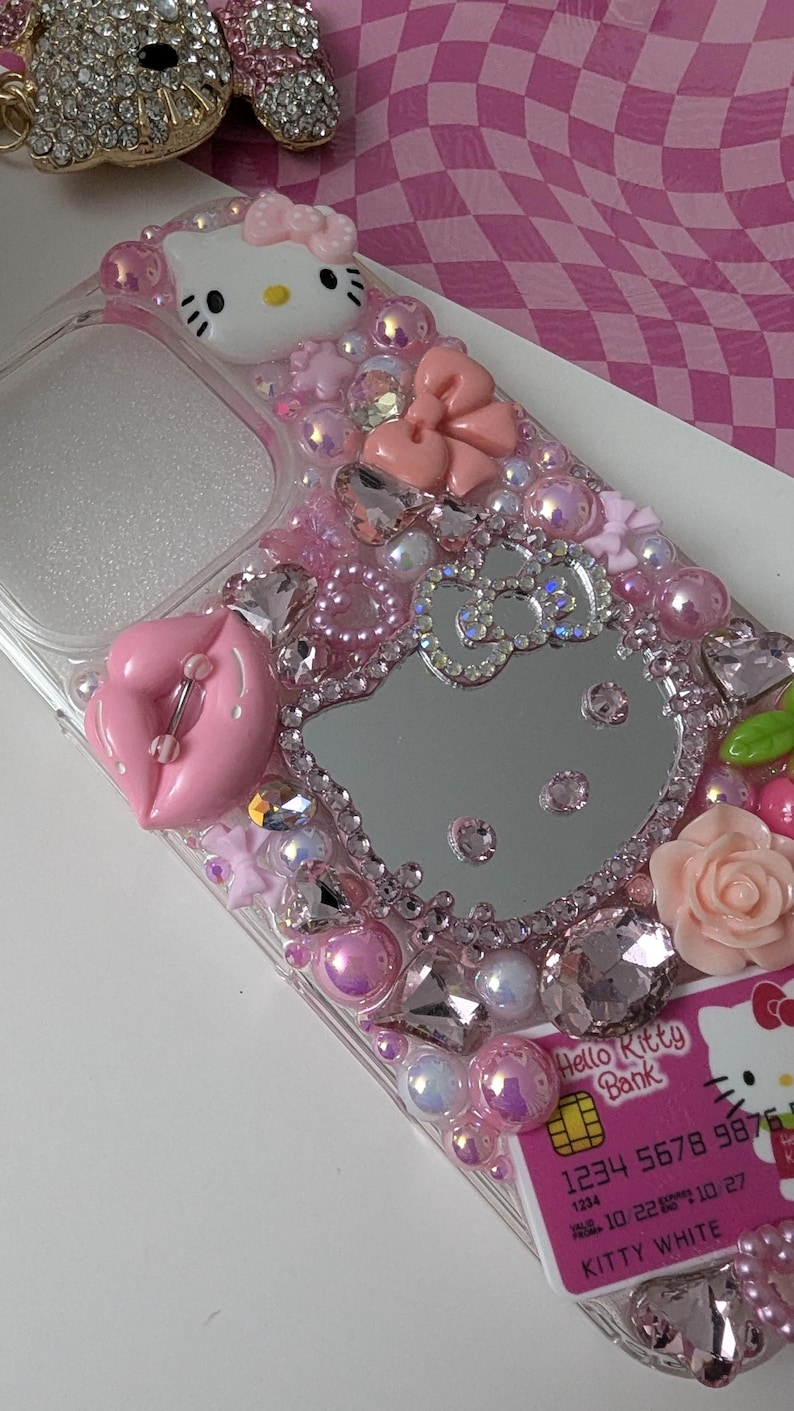 May include: A clear phone case bedazzled with pink and clear rhinestones, pearls, and charms. The case features a Hello Kitty design, a pink bow, and a pink lip charm. A Hello Kitty Bank credit card is visible.