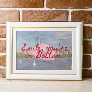 May include: Framed art print with a gold border, featuring a Boston cityscape with sailboats. The artwork includes the text "Smile, you're in Boston" in red script. A white vase with yellow flowers sits to the right.