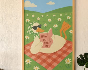 Op de afbeelding: Een ingelijste kunstprint met een cartoon eend met een zonnebril en een hoofddoek, die een boek leest met de titel "HOW TO TRICK MEN". De eend ligt op een rood-wit geruite deken in een madeliefjesveld.