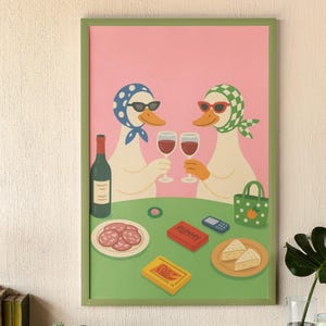 Op de afbeelding: Een ingelijste kunstprint met twee antropomorfe eenden met zonnebrillen en hoofddoeken die toosten met wijnglazen. De sc&egrave;ne omvat een fles wijn, snacks en een Rummy-spel, geplaatst op een groene tafel tegen een roze achtergrond.