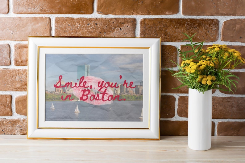 May include: Framed art print with a gold border, featuring a Boston cityscape with sailboats. The artwork includes the text "Smile, you're in Boston" in red script. A white vase with yellow flowers sits to the right.