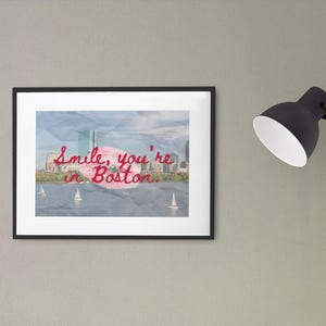 May include: A framed print featuring a Boston cityscape with sailboats on the water. The artwork has the text "Smile, you're in Boston" in red script. The frame is black, and the print is displayed on a gray wall.
