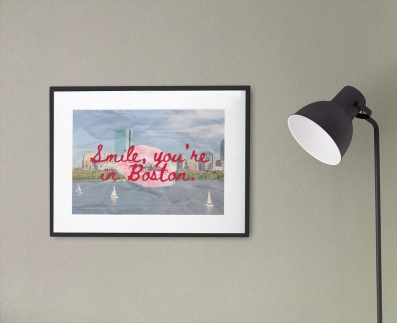 May include: A framed print featuring a Boston cityscape with sailboats on the water. The artwork has the text "Smile, you're in Boston" in red script. The frame is black, and the print is displayed on a gray wall.