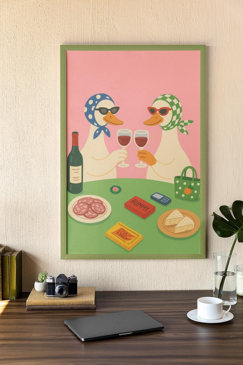 Op de afbeelding: Een ingelijste kunstprint met twee antropomorfe eenden met zonnebrillen en hoofddoeken die toosten met wijnglazen. De sc&egrave;ne omvat een fles wijn, snacks en een Rummy-spel, geplaatst op een groene tafel tegen een roze achtergrond.