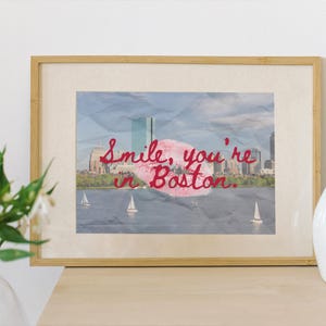 May include: Framed print featuring a Boston cityscape with sailboats on the water. The image has a textured, crumpled paper effect. Red script reads "Smile, you're in Boston." A white vase and flowers are in the foreground.