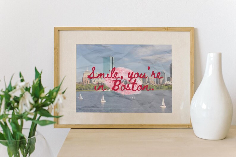 May include: Framed print featuring a Boston cityscape with sailboats on the water. The image has a textured, crumpled paper effect. Red script reads "Smile, you're in Boston." A white vase and flowers are in the foreground.