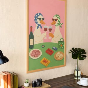 Op de afbeelding: Een ingelijste kunstprint met twee antropomorfe ganzen met zonnebrillen en hoofddoeken, die toosten met wijnglazen. Een tafel is gedekt met eten, een fles wijn en een Rummy-spel. De achtergrond is roze en groen.