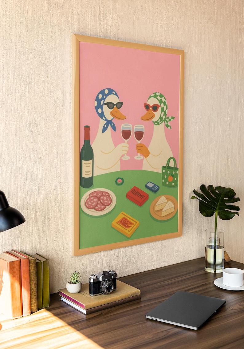 Op de afbeelding: Een ingelijste kunstprint met twee antropomorfe ganzen met zonnebrillen en hoofddoeken, die toosten met wijnglazen. Een tafel is gedekt met eten, een fles wijn en een Rummy-spel. De achtergrond is roze en groen.