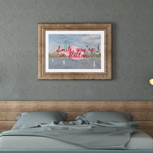 May include: A framed print with the text "Smile, you're in Boston" in red script. The image inside the frame depicts a cityscape with sailboats on the water. The frame is a light brown color and is hanging above a bed.