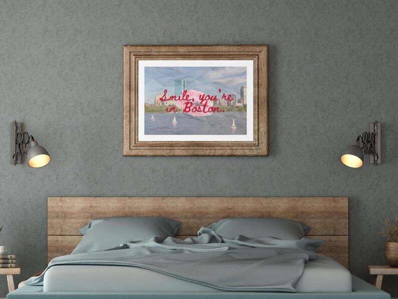 May include: A framed print with the text "Smile, you're in Boston" in red script. The image inside the frame depicts a cityscape with sailboats on the water. The frame is a light brown color and is hanging above a bed.