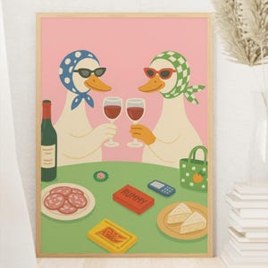 Op de afbeelding: Een ingelijste kunstprint met twee antropomorfe eenden met zonnebrillen en hoofddoeken, die toosten met wijnglazen. Een tafel is gedekt met eten, een fles wijn en een Rummy-spel. De achtergrond is roze.