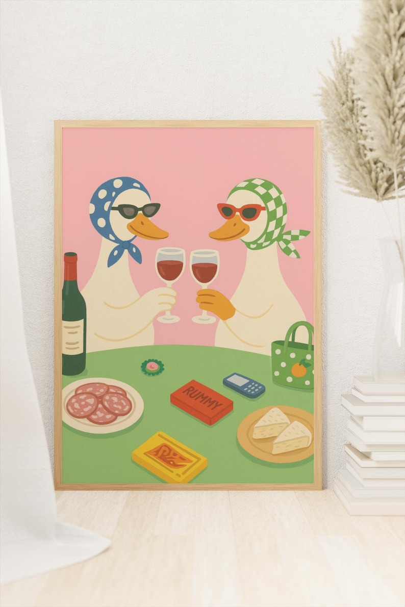 Op de afbeelding: Een ingelijste kunstprint met twee antropomorfe eenden met zonnebrillen en hoofddoeken, die toosten met wijnglazen. Een tafel is gedekt met eten, een fles wijn en een Rummy-spel. De achtergrond is roze.