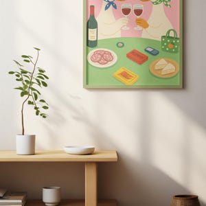 Op de afbeelding: Een ingelijste illustratie toont twee ganzen met zonnebrillen en hoofddoeken, proostend met wijnglazen. Een tafel is gedekt met eten, waaronder salami, kaas en een speldoos. De achtergrond is roze en de lijst is groen.