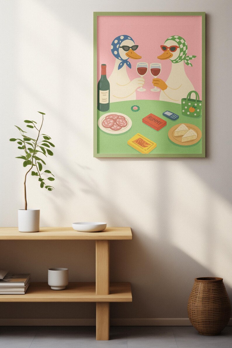 Op de afbeelding: Een ingelijste illustratie toont twee ganzen met zonnebrillen en hoofddoeken, proostend met wijnglazen. Een tafel is gedekt met eten, waaronder salami, kaas en een speldoos. De achtergrond is roze en de lijst is groen.