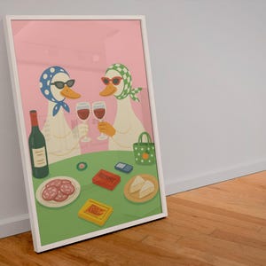Op de afbeelding: Ingelijste kunstprint met twee antropomorfe eenden met zonnebrillen en hoofddoeken, proostend met wijnglazen. Een tafel toont een fles wijn, snacks en een Rummy-spel. De achtergrond is roze.