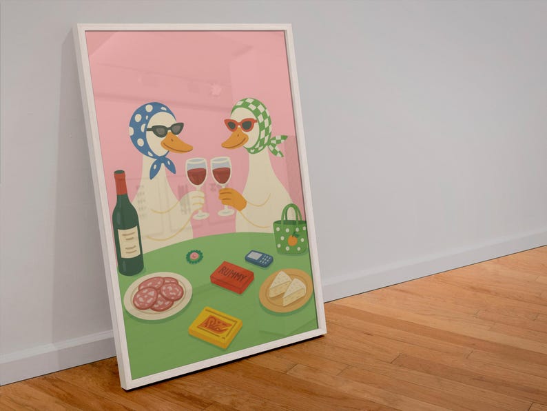 Op de afbeelding: Ingelijste kunstprint met twee antropomorfe eenden met zonnebrillen en hoofddoeken, proostend met wijnglazen. Een tafel toont een fles wijn, snacks en een Rummy-spel. De achtergrond is roze.