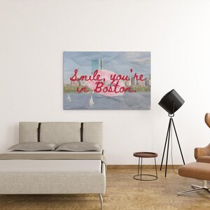 May include: A canvas print with the text "Smile, you're in Boston" in red script over a watercolor image of the Boston skyline. The print hangs above a bed with a light beige headboard and a modern brown leather chair.