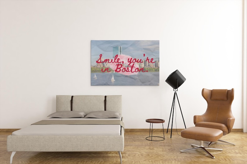 May include: A canvas print with the text "Smile, you're in Boston" in red script over a watercolor image of the Boston skyline. The print hangs above a bed with a light beige headboard and a modern brown leather chair.