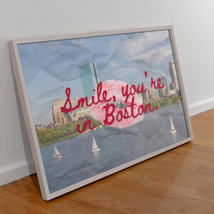 May include: Framed art print featuring a Boston cityscape with sailboats on the water. The artwork includes the text "Smile, you're in Boston" in red script over a pink lip print, set against a blue sky background.