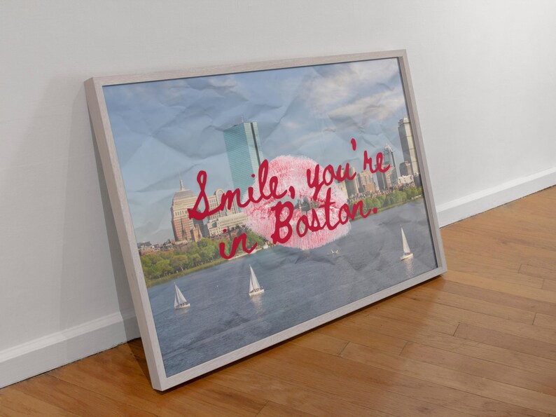 May include: Framed art print featuring a Boston cityscape with sailboats on the water. The artwork includes the text "Smile, you're in Boston" in red script over a pink lip print, set against a blue sky background.