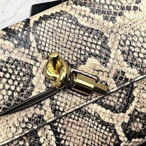 May include: Close-up of a snakeskin-patterned handbag. The bag features a gold-colored knot closure and a gold-colored rectangular clasp. The snakeskin pattern is a mix of black, white, and tan colors.