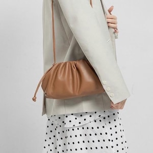 Puede incluir: Un bolso bandolera de cuero color canela con un diseño fruncido. El bolso tiene una correa larga y fina y un cierre de cordón. El bolso se lleva sobre una blazer color crema y una falda de lunares blancos y negros.